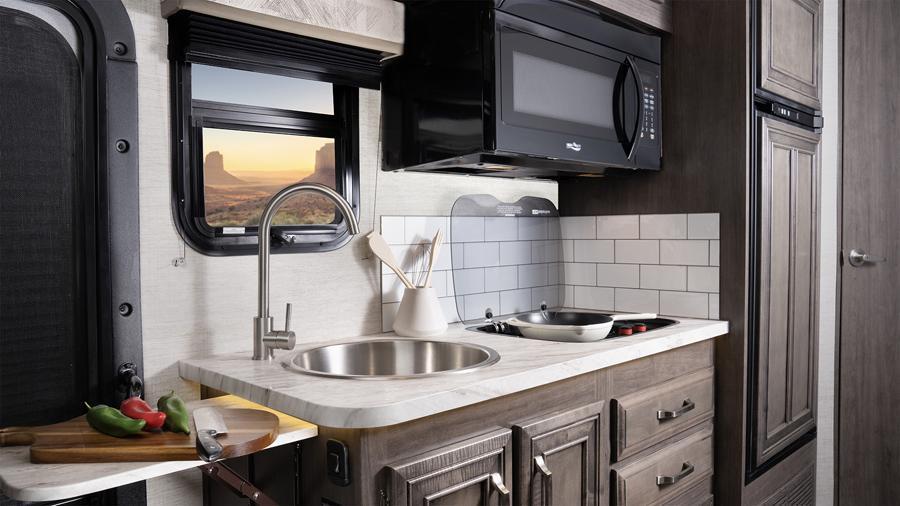 Qwest SE 24L Kitchen | The kitchen in the Qwest SE 24L has a 10-cubic-foot 12V refrigerator, a 2-burner, drop-in cooktop with flush cover, a convection microwave, a stainless steel sink with a wooden cover/cutting board, LED-lit pressed countertops, a flip-up countertop extension (select models) and a decorative backsplash.